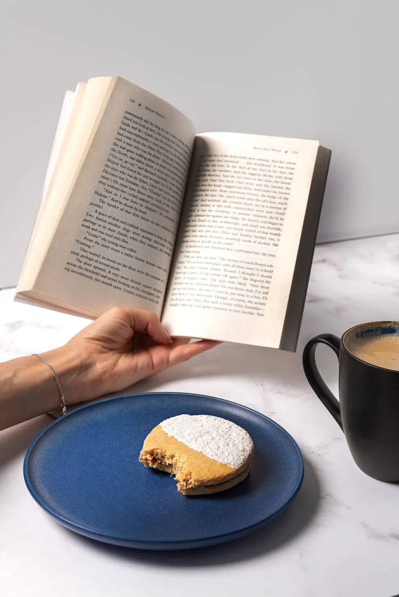 Alfajor Gigante mordido en plato azul, junto a un libro y una taza de café