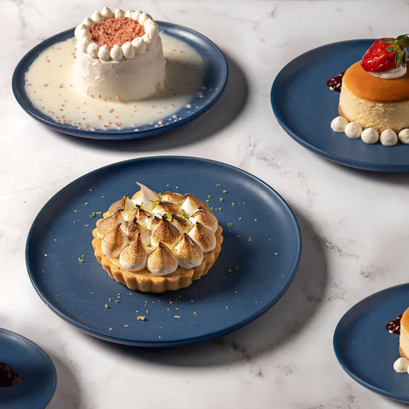 Tres Leches de Piñón, NY Cheesecake con Fresas y Key Lime Pie en platos azules sobre mármol, vista cenital